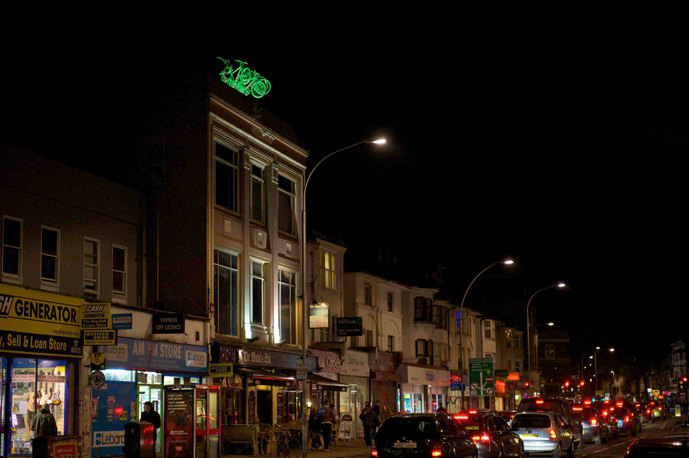 5 brightly lit, green bikes leap into a night sky, from the roof of a tall building, over a shopping street