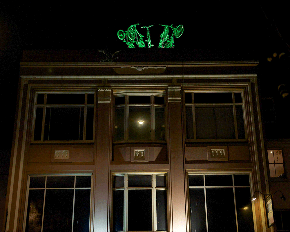 5 brightly lit, green painted bikes leap from the roof of an art deco building